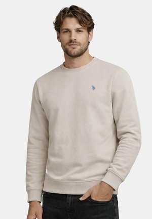 Man wearing a beige crewneck sweatshirt with a small blue logo on the chest, standing with one hand in pocket against a plain background.
