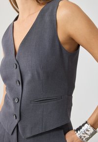 Pelēks piegrieztais veste ar V-veida kakla izgriezumu, sešiem pogām un krūšu kabatu. Materiāls ir gluds, ar gludu tekstūru un strukturētu dizainu.