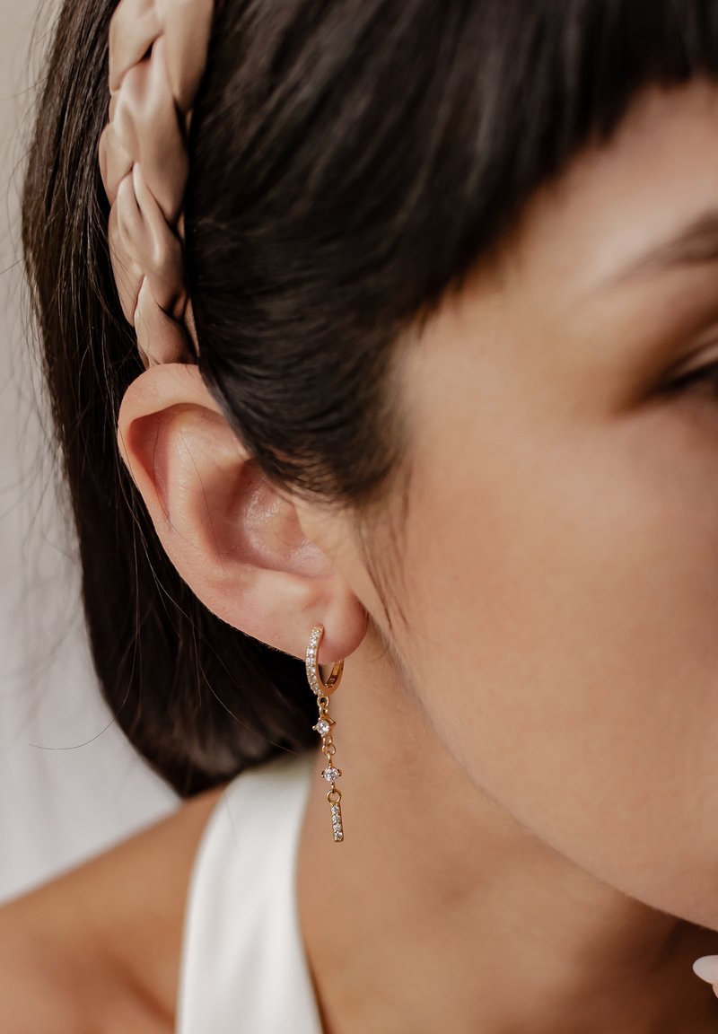 Goldene Creolen mit einer herabhängenden Kette, die klare Edelsteine zeigt. Das Design umfasst eine Mischung aus kleinen und größeren Steinen für visuelles Interesse.