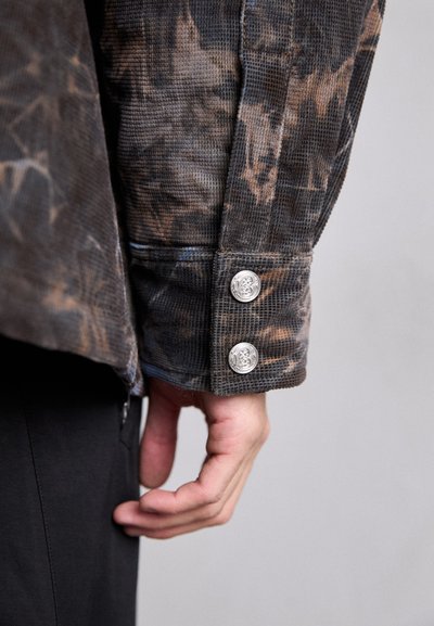 Brown corduroy shirt with a textured pattern, featuring silver buttons at the cuff. Close-up showing the arm and hand positioned near the body.