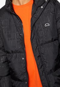 Czarna pikowana kurtka z błyszczącym wykończeniem, ozdobiona subtelnym wytłoczonym wzorem. Z przodu zapinana na zamek błyskawiczny, z małą naszywką z logo. Pod spodem pomarańczowy t-shirt.