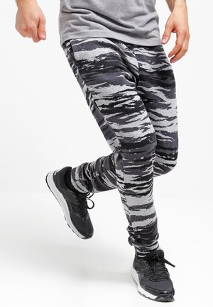 Person trägt graue und schwarze tigergestreifte Hose und schwarze Sneakers, beim Mittelschritt vor einem schlichten weißen Hintergrund.