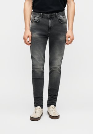 Man wearing slim-fit faded black jeans and off-white sneakers, standing against a plain light background.