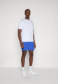 Mann som står iført en enkel hvit kortermet skjorte, blå shorts, hvite sokker og hvite sneakers foran en enkel lys bakgrunn.
