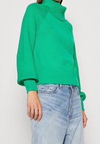 Pull vert avec un col montant, des manches en maille texturée, des poignets côtelés et une coupe décontractée, associé à un jean bleu clair.