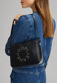 Borsa a tracolla in pelle nera con una superficie liscia, caratterizzata da borchie argentate disposte in un motivo circolare e una chiusura con zip. Include una tracolla.