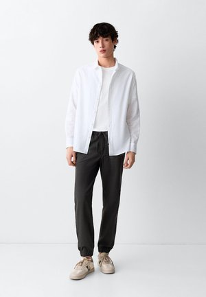 Junger Mann mit weißem Hemd über weißem T-Shirt, schwarzen Jogginghosen und beigen Sneakers, steht vor einem einfarbigen weißen Hintergrund.