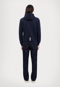 Mørkeblå hettegenser med en avslappet passform, laget av en bomullsblanding, og med hvite striper på baksiden, kombinert med mørke denimjeans.