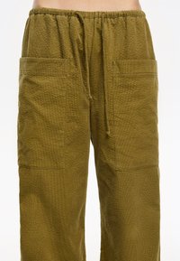 Pantalones de pierna ancha de color verde oliva, con textura, y con una cinturilla elástica y cordón; cuentan con dos grandes bolsillos delanteros tipo parche.