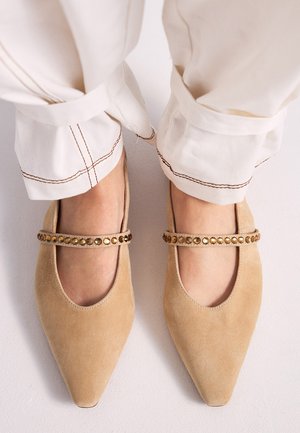 Pies con zapatos planos de ante beige con punta y tiras con tachuelas, combinados con pantalones crema con costuras marrones en los puños.