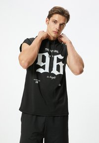 Schwarze ärmellose Kapuzenjacke mit einem auffälligen weißen Aufdruck, der "96" und "DIE ZUKUNFT ist hell." zeigt. Hergestellt aus glattem, leichtem Stoff.