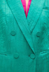 Blazer turquesa de doble botonadura con un tejido texturizado, que presenta botones redondos a juego y dos bolsillos laterales. Camisa rosa visible debajo.