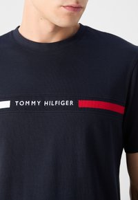 Férfi, aki tengerészkék pólót visel, amelyen a „Tommy Hilfiger” felirat fehér és piros vízszintes csíkok között helyezkedik el a mellkasán.