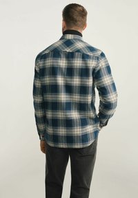 Niebieska w kratę flanelowa koszula z długimi rękawami, z ostrym kołnierzykiem i zaokrąglonym dołem, noszona z ciemnoszarymi spodniami.