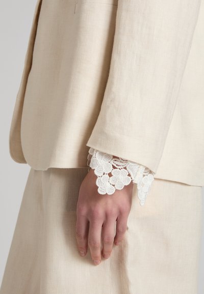 Hand rustend langs het lichaam, gekleed in een crèmekleurig pak met een bloemrijke kanten manchetdetail zichtbaar onder de mouw.