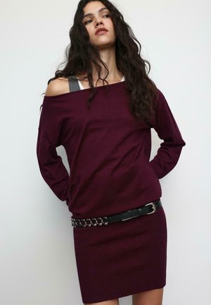 Bordo pulover otvorenih ramena, pletene teksture, s pripijenom rebrastom suknjom i crnim kožnim remenom s naglašenim srebrnim lancima.