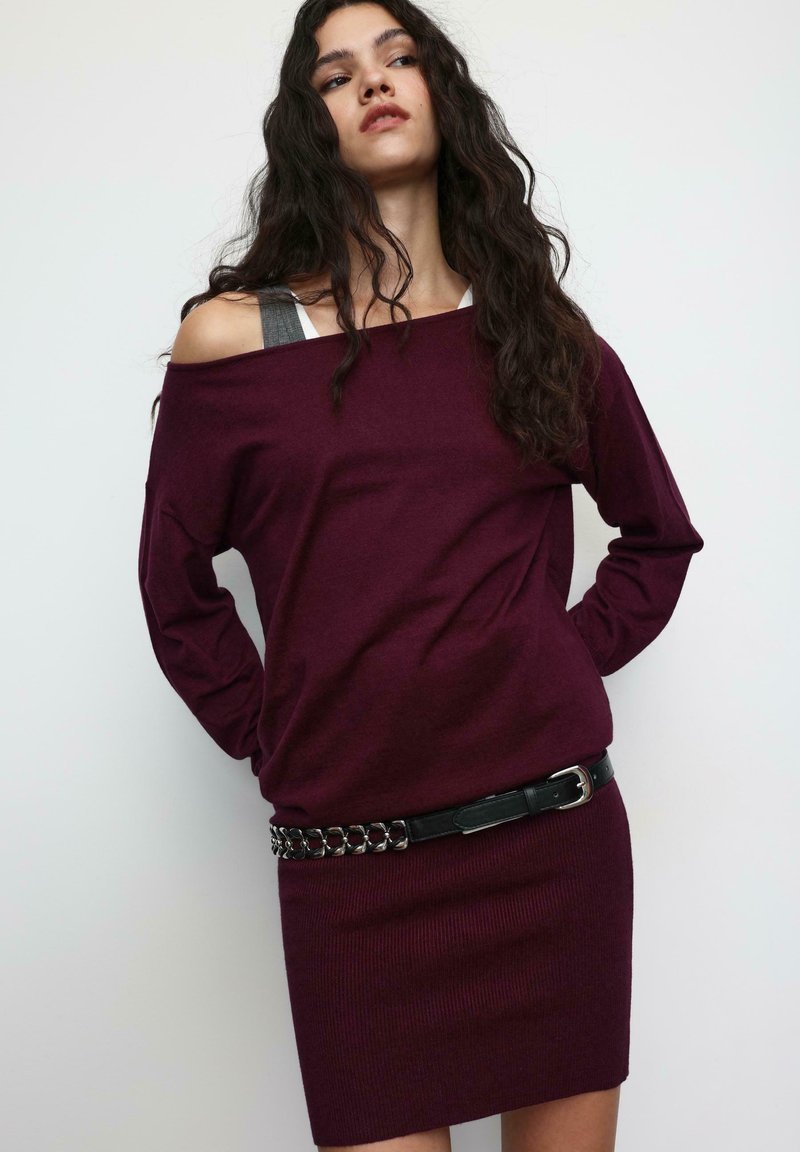 Bordeauxrode trui met blote schouders, gebreide textuur, met aansluitende geribbelde rok en zwarte leren riem met zilveren kettingaccenten.