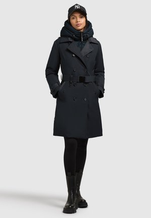 Trench-coat bleu marine foncé avec un design à double boutonnage, taille ceinturée et capuche ajustable sur une doudoune noire, associé à des leggings noirs et des bottes.