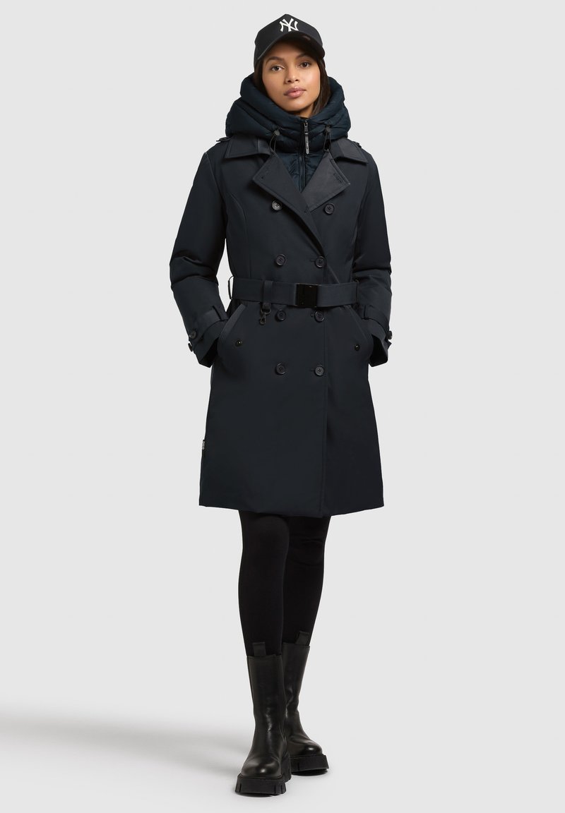 Trench coat blu scuro con design doppiopetto, vita con cintura e cappuccio regolabile indossato su un giubbino imbottito nero, abbinato a leggings neri e stivali.