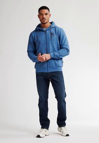 Felpa con cappuccio blu con zip e coulisse, realizzata in tessuto strutturato. Abbinata a jeans scuri in denim e scarpe sportive bianche.