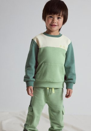 Young boy smiling, wearing a green and cream long-sleeve sweatshirt and matching light green pants with side pockets.
