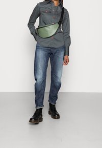 Grüner, glatt strukturierter Bauchtasche, getragen über einem grauen Shirt mit roten Knöpfen, kombiniert mit blauen Jeans und schwarzen Schnürstiefeln.