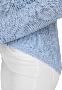 Maglione a maglia blu chiaro con trama allentata, orlo a coste e fondo arrotondato. Abbinato a pantaloni in cotone bianchi, che mostrano i dettagli laterali.
