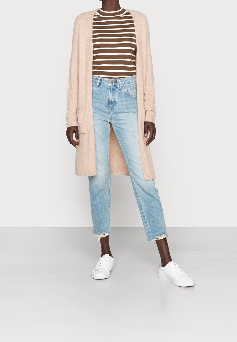 Beige kavaj med öppen front och fickor, randig brun- och vit långärmad tröja, ljusblå slitna jeans och vita sneakers.