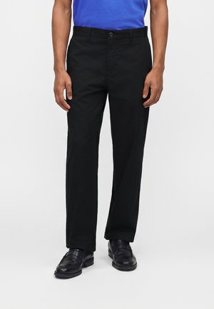 Homme portant un pantalon noir à jambes droites, des chaussures en cuir noires et une chemise bleue sur un fond clair uni.