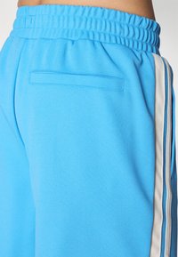 Calças de jogging azul céu com um tecido texturado, cintura elástica e um único bolso traseiro. Apresenta riscas laterais brancas para contraste.