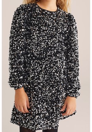 Niño con un vestido negro de manga larga cubierto de lentejuelas plateadas densamente distribuidas, de pie contra un fondo liso.