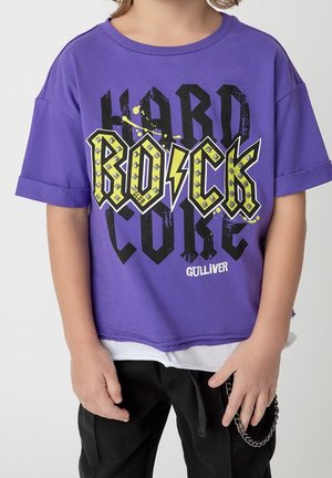 Dziecko ubrane w fioletowy T-shirt z krótkim rękawem z napisami „Rock Hard Core” i „Gulliver”, czarne spodnie oraz łańcuch jako dodatek zwisający z talii.