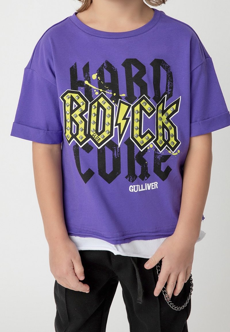 Barn iført en lilla T-shirt med korte ærmer med teksten "Rock Hard Core" og "Gulliver", sorte bukser og en kæde ved hoften.