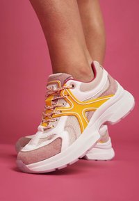 Růžové a bílé sportovní boty s síťovinovým svrškem, semišovými akcenty, žlutými detaily a texturovanými tkaničkami. Tlustá bílá podrážka s průhlednou částí.
