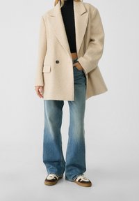 Beige, textuur oversized jas met grote revers en enkele knoopsluiting, gecombineerd met lichtblauwe flared jeans en bruine sneakers.