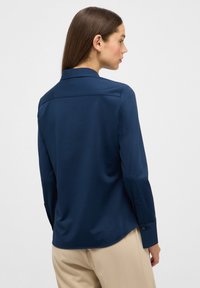 Camicia abbottonata blu navy con una texture liscia, maniche lunghe e colletto classico. Presenta un orlo curvo e polsini abbottonati.