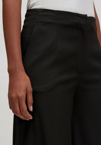 Schwarze maßgeschneiderte Hose mit glatter Stoffoberfläche, ausgestattet mit vorderen Falten und klassischer Passform, präsentiert mit einer Hand, die auf dem Stoff ruht.