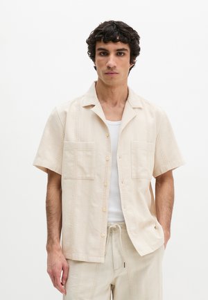 Junger Mann mit lockigem dunklem Haar, der ein beiges Kurzarm-Hemd über einem weißen Tanktop und passende beige Hosen trägt.