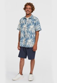 Camisa de manga corta con un patrón de hojas tropicales en azul y blanco; combinada con pantalones cortos azul marino. Las zapatillas son blancas con detalles en negro.