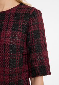 Haut à manches courtes rouge et noir en tartan, en tissu texturé avec un ourlet légèrement effiloché. Accents scintillants tissés tout au long.