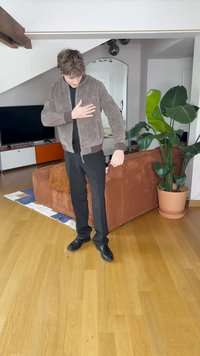 Καφέ σουέτ bomber jacket με ριγέ μανσέτες και στρίφωμα, φορεμένο πάνω από μαύρο πουκάμισο. Συνδυασμένο με μαύρο παντελόνι και παπούτσια. Εσωτερικός χώρος.