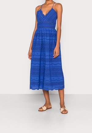 Frau in einem blauen ärmellosen Midikleid mit Lochmuster und gewelltem Saum, kombiniert mit beigen flachen Sandalen.