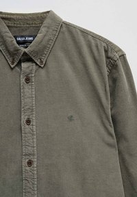 Camisa de pana verde con collar abotonado, botones marrones y un pequeño logotipo bordado en el lado izquierdo. Diseño de corte regular.