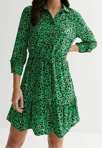 Abito verde con bottoni, caratterizzato da una stampa leopardata, vita arricciata e maniche lunghe, con una gonna a strati e tessuto leggero.
