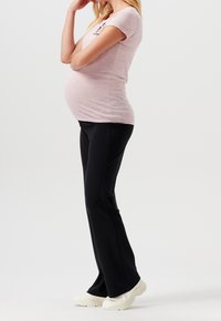 Těhotenský outfit zahrnující světle růžové tričko s krátkým rukávem a grafickým designem, doplněné černými elastickými kalhotami a bílými teniskami.