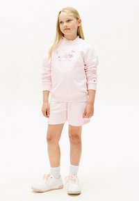 Felpa di cotone rosa con scollo arricciato e testo del logo, abbinata a pantaloni corti rosa coordinati. Sneakers bianche con dettagli rosa e calzini alti.