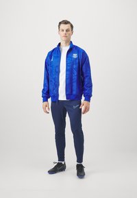 Blauwe sportjack met textuurpatronen, ritssluiting aan de voorkant en logo. Gecombineerd met marineblauwe broek en zwarte voetbalschoenen met blauwe accenten.