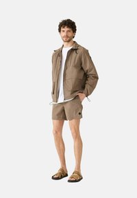 Lichte bruin jas met een rits en zakken, gecombineerd met bijpassende shorts. Gedragen met bruine sandalen met banden, staand tegen een effen achtergrond.