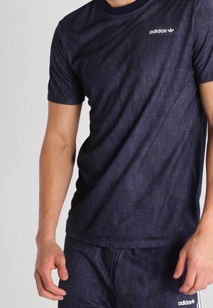 Mand iført mørkeblå Adidas t-shirt og matchende bukser, stående med armene afslappet langs siderne mod en ensfarvet baggrund.