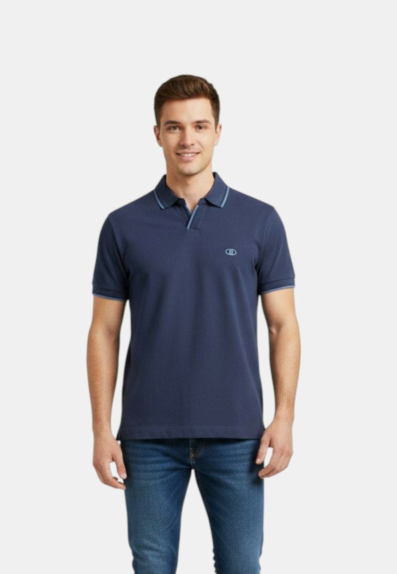 Homme portant un polo bleu marine à manches courtes et un jean bleu, debout, souriant, sur un fond blanc uni.